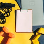 Flat lay of fitness equipment including dumbbells, jump rope, and clipboard on a yellow background.